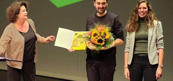 Johannes Hebsacker bei der Preisübergabe des Klaus Zehelein Preises 2022, in der Hand einen Blumenstrauss, rechts von ihm Antonia Leitgeb (Stellv. Studiengangsleiterin Studiengang Dramaturgie Theaterakademie August Everding), links Almut Wagner (Chefdramaturgin Residenztheater München)