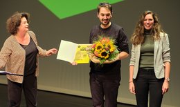 Johannes Hebsacker bei der Preisübergabe des Klaus Zehelein Preises 2022, in der Hand einen Blumenstrauss, rechts von ihm Antonia Leitgeb (Stellv. Studiengangsleiterin Studiengang Dramaturgie Theaterakademie August Everding), links Almut Wagner (Chefdramaturgin Residenztheater München)
