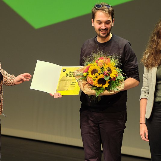Johannes Hebsacker bei der Preisübergabe des Klaus Zehelein Preises 2022, in der Hand einen Blumenstrauss, rechts von ihm Antonia Leitgeb (Stellv. Studiengangsleiterin Studiengang Dramaturgie Theaterakademie August Everding), links Almut Wagner (Chefdramaturgin Residenztheater München)